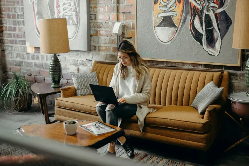 A woman working on a laptop while seated on a sofa in a bright, brick-walled workspace